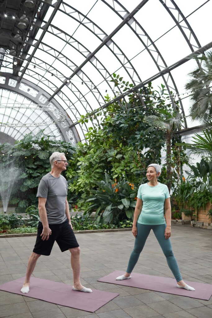 Elderly couple practicing yoga in a lush greenhouse, promoting health and wellness.